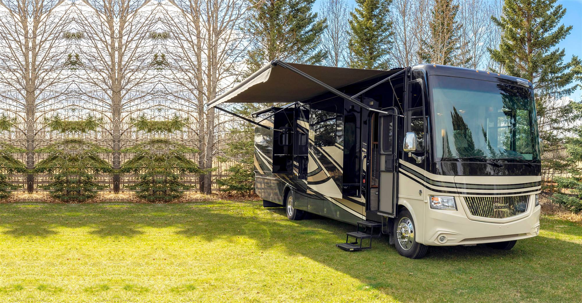 A Class A motorhome with its awning out, parked on the grass among pine trees