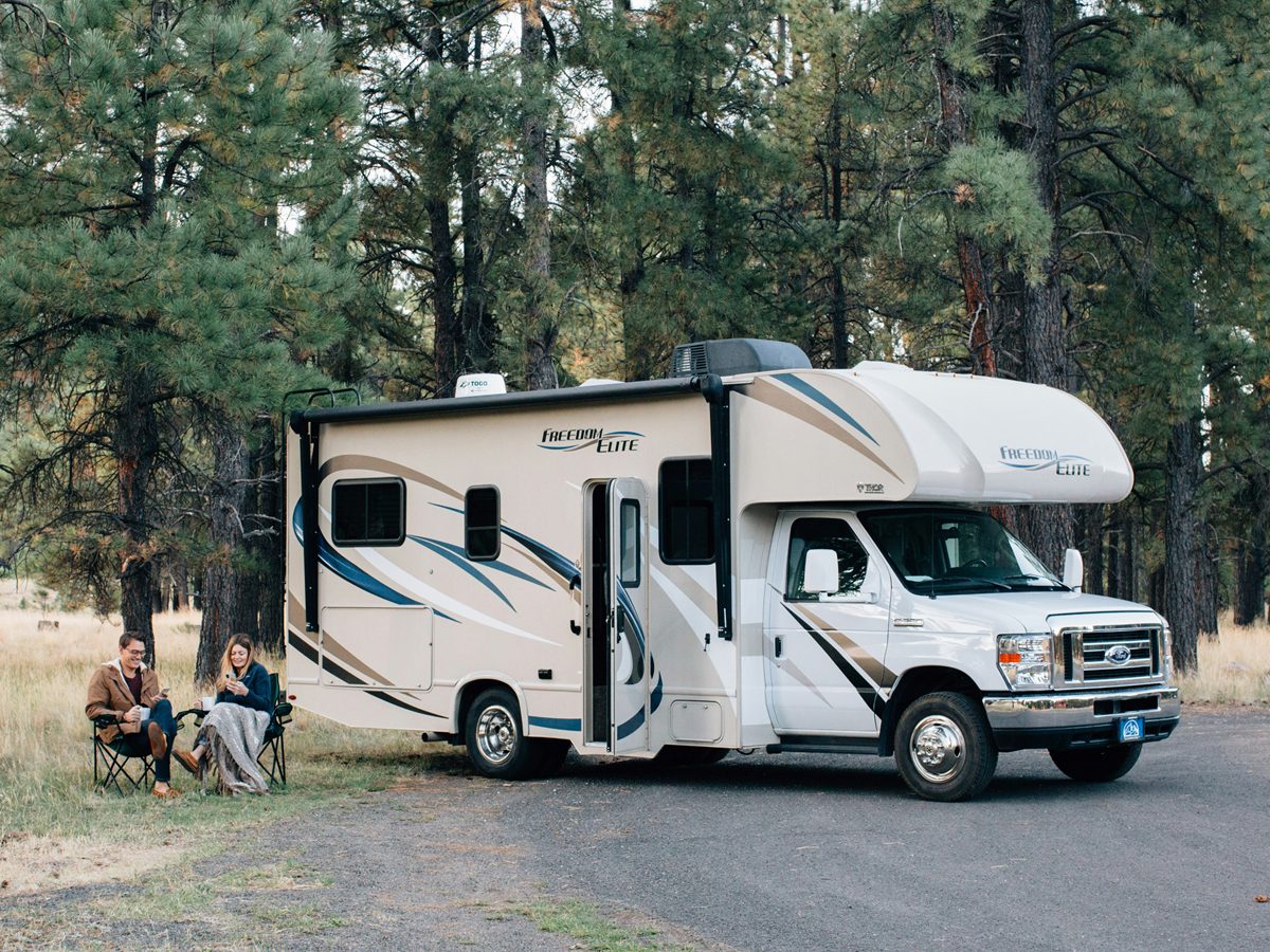 A Class C motorhome parked among pine trees while its owners relax beside it